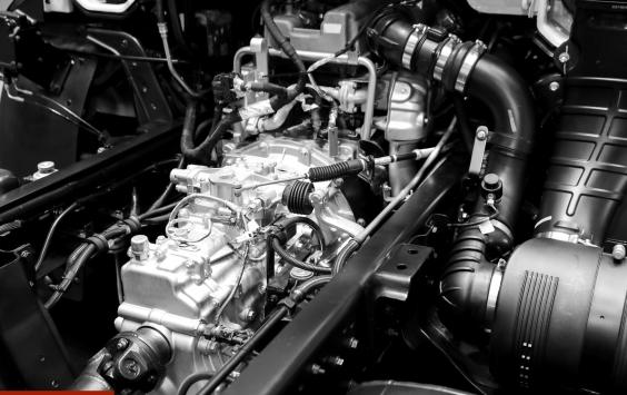 Close-up of a vehicle engine bay showing the engine, pipework and wiring, in black and white.